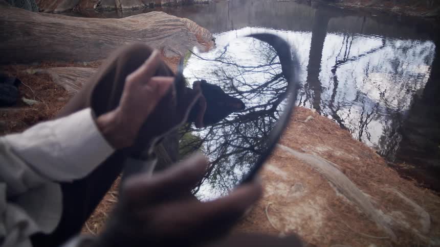 An elderly man sits on a large rock in front of a river and looks in a mirror