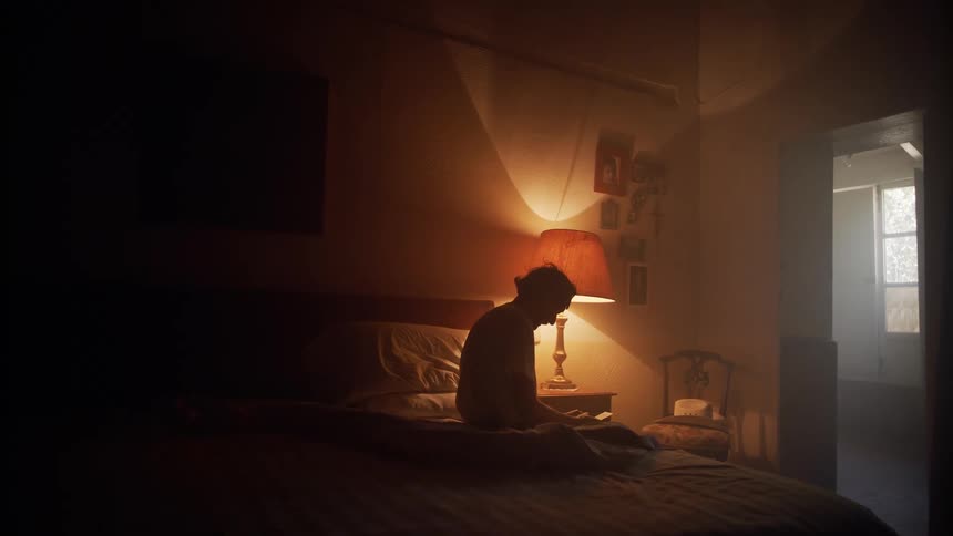 A person is backlit by a lamp while sitting in a dark bedroom