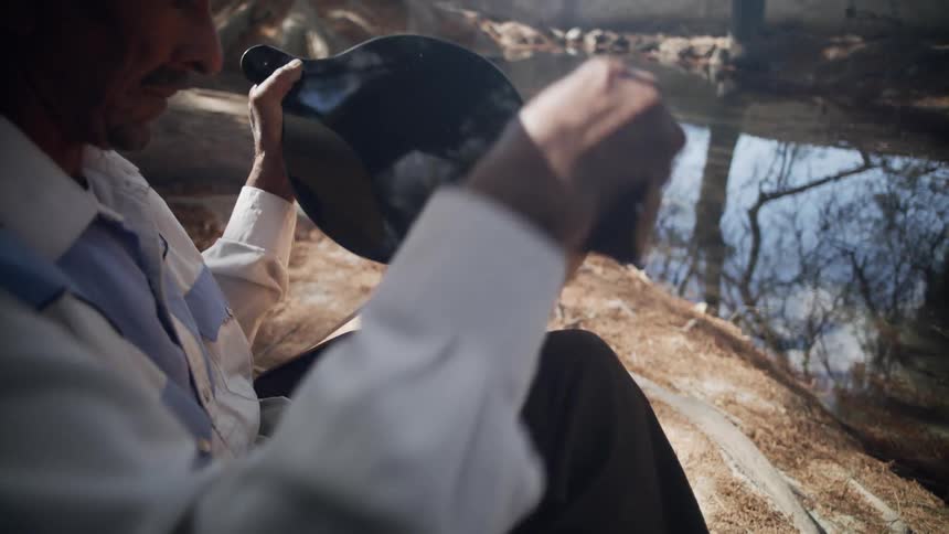 Man wearing straw hat unwraps mirrored disk that reflects trees similarly to the nearby still water