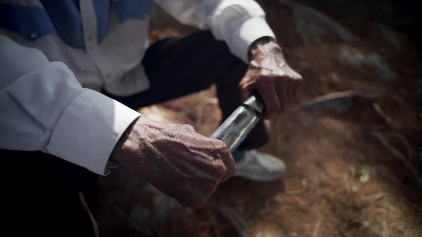 Elderly man sitting in the ground holds a shiny knife in his hands