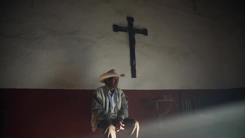 A man leans against the wall of a church beneath a large cross