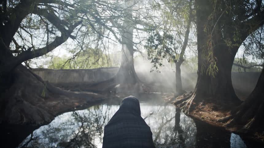 A person covered in a plaid scarf sits in front of a small pond in a forest