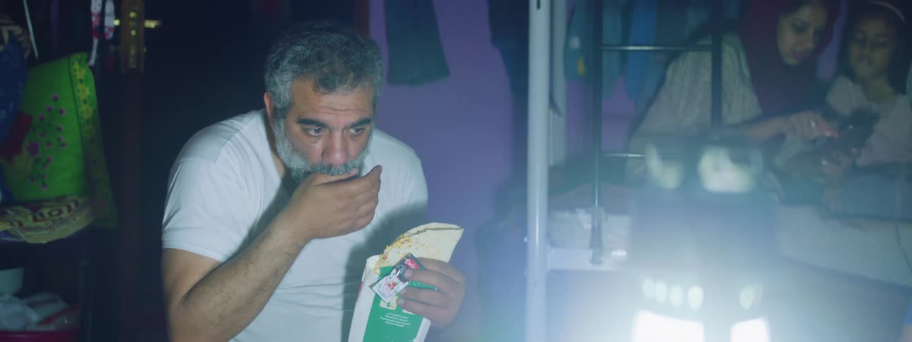 A young woman sits down in front of an electric lantern in a dark room next to an older man trying to open a condiment packet for his burrito