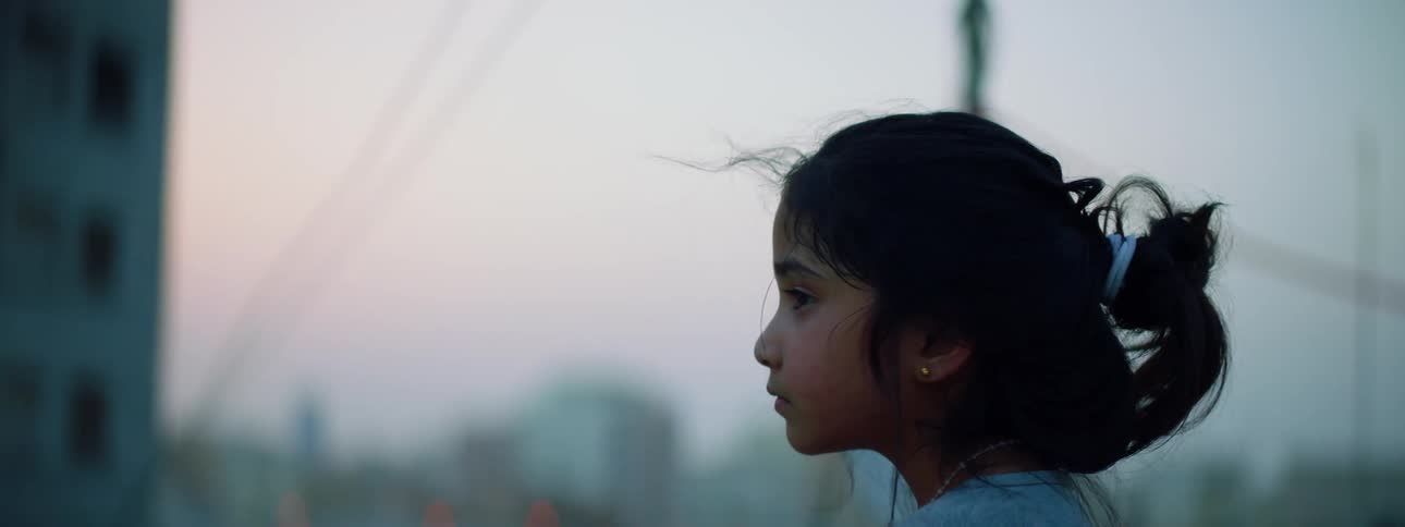 A little girl climbs onto the railing on the edge of a rooftop and looks out over the city