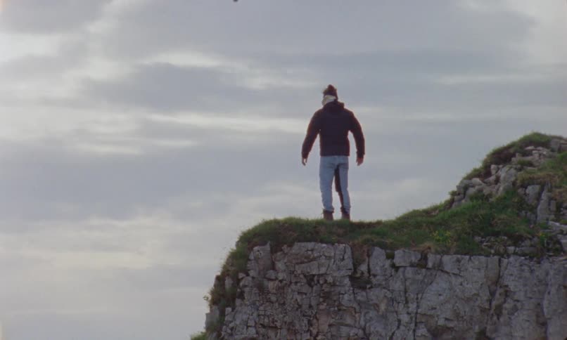 Man with a coat jeans and a pair of boots stands at the edge of a cliff on a cloudy day