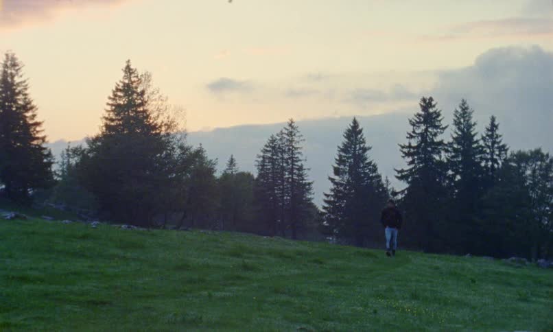 Man with blue pants a pair of boots a black coat and a black headband walks down a grass hill at dusk