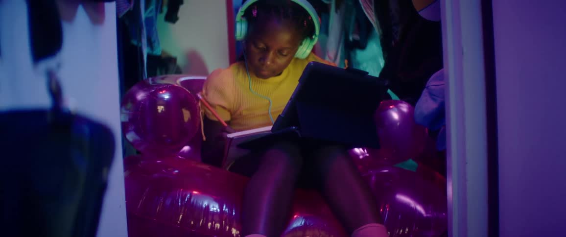 A girl sitting on a bloated couch writing on a book with a pencil and watching a laptop