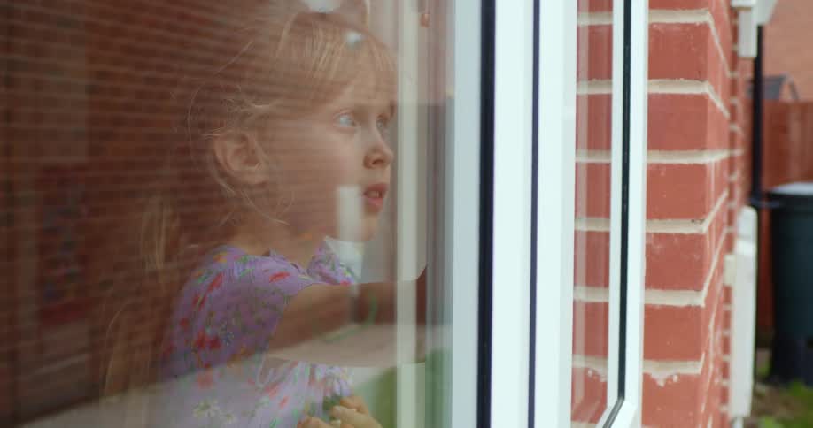 A girl in a blue floral dress draws on the inside of a window