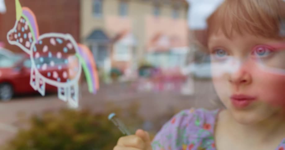 Blonde girl with blue eyes draws an unicorn with colorful markers on the window of a house