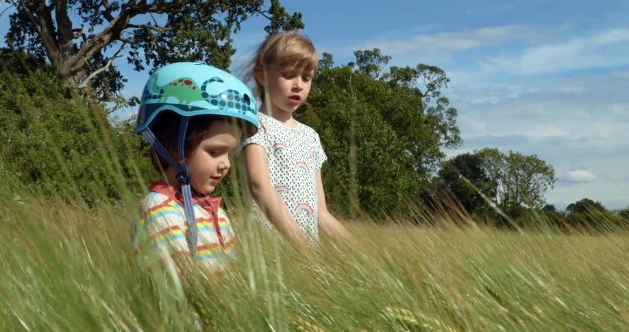 Blonde girl and a boy with a blue helmet stands in tall grass on a sunny day