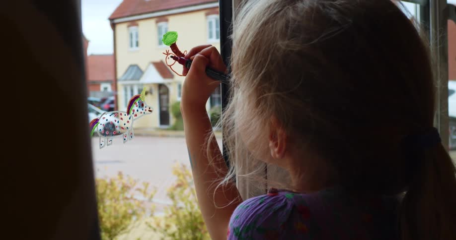 A little girl draws on a window in her home