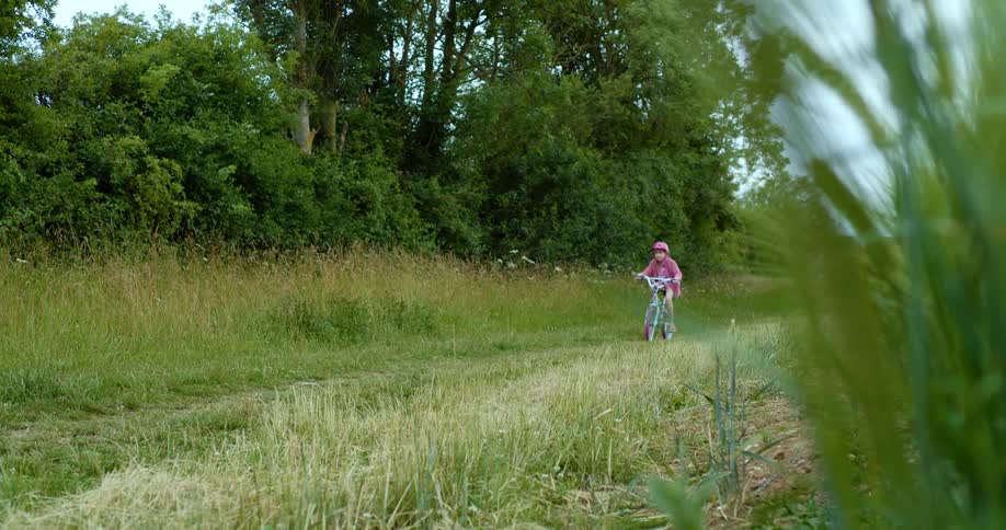 Girl with a pink helmet and sweater rides a bicycle on a dirt road in the countryside