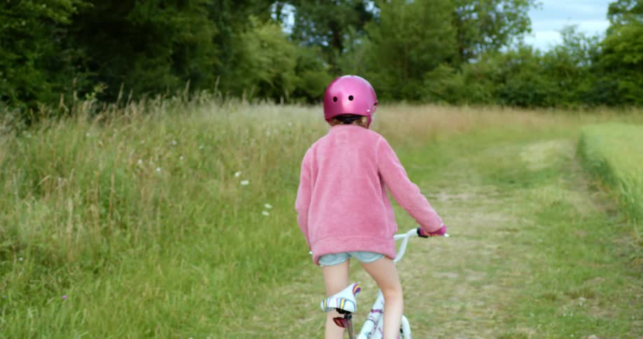 Girl with a pink helmet and sweater rides a bicycle on a dirt road in the countryside