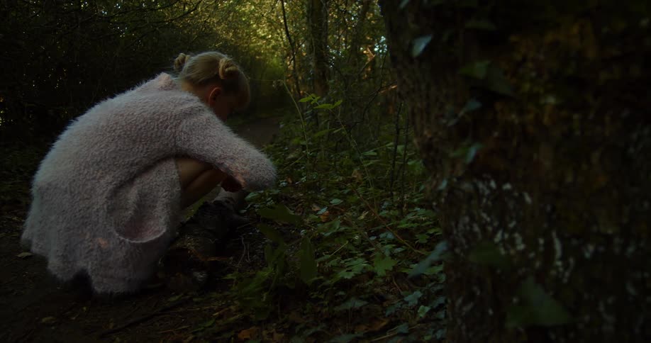 A young girl crouches down as she picks plants on the ground in a forest