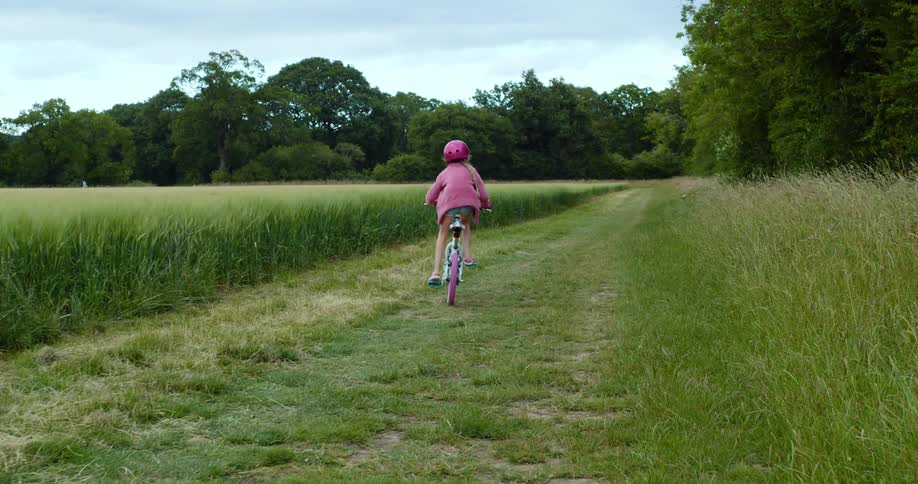A young girl in a pink jacket rides a pink bicycle through a path in a field