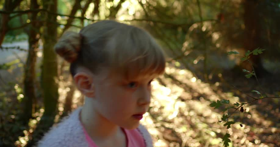 Blonde girl with a grey pullover and a pink shirt walks on the brown leaves as the sun shines bright in the forest
