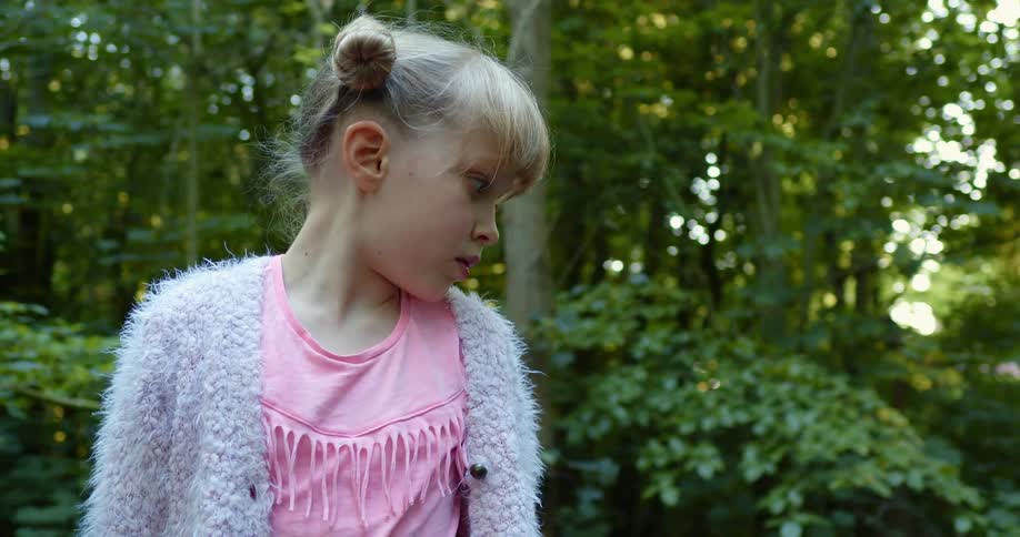Girl wearing pink sweater and shirt stands looking around the forest
