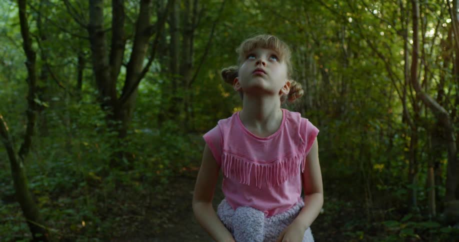 A little girl looks all around her as she waves through a thick forest during the day