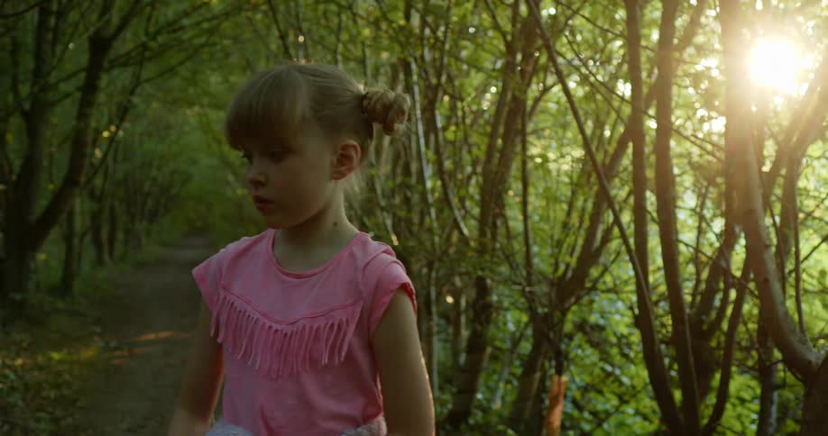 A young girl in a pink shirt holds a stick while walking on a dirt path in a forest