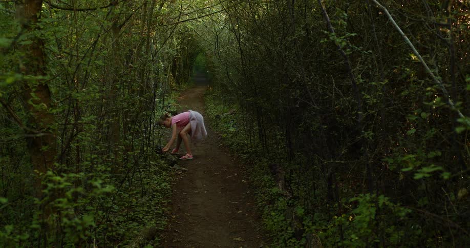 Girl walks along narrow dirt trail through woods stopping to pickup log