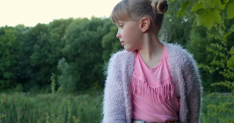 Young girl in a cardigan sweater stands in a field next to a forest