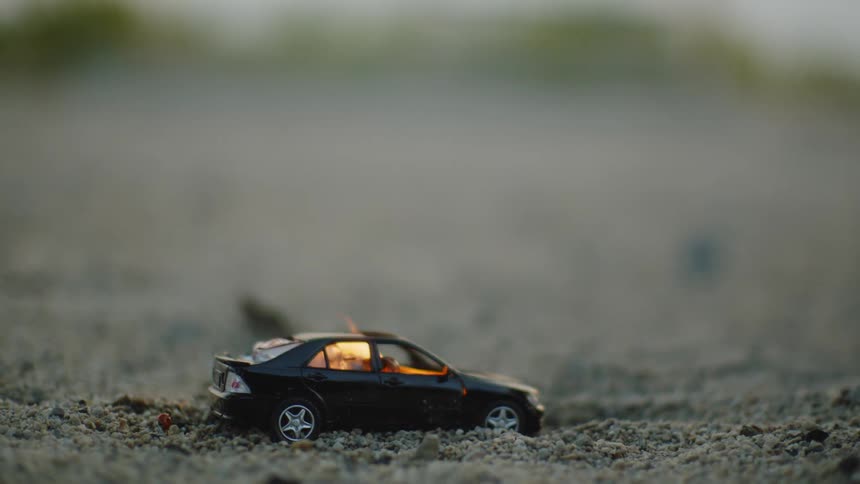 Flames engulf a toy car as it lies in the soil outdoors