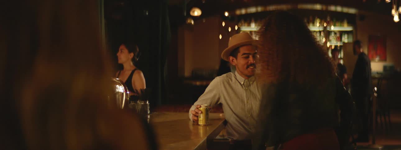Woman approaches a man sitting at the bar and sits next to him as another woman watches