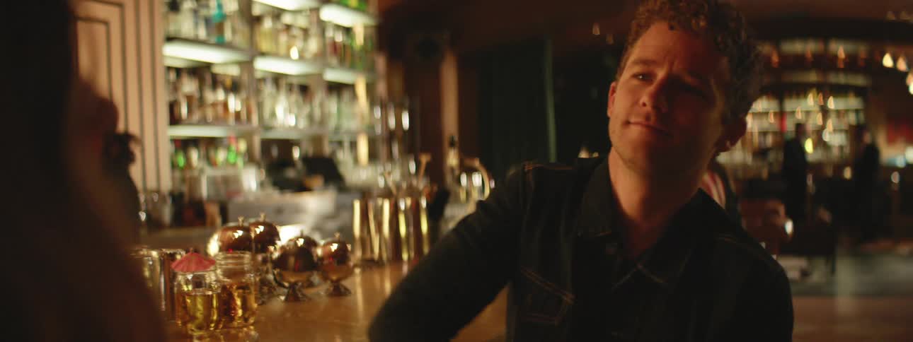 Man with a denim jacket sitting at bar smiles looks at a woman
