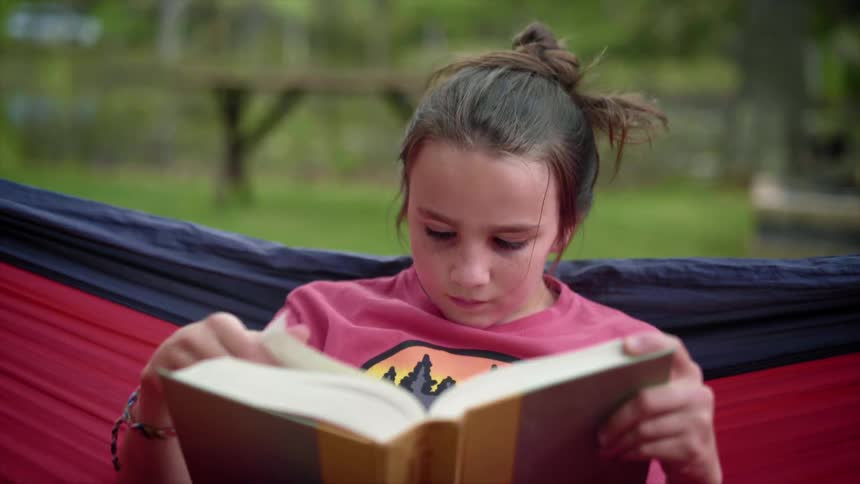 A girl reads a book while rocking in a hammock outdoors