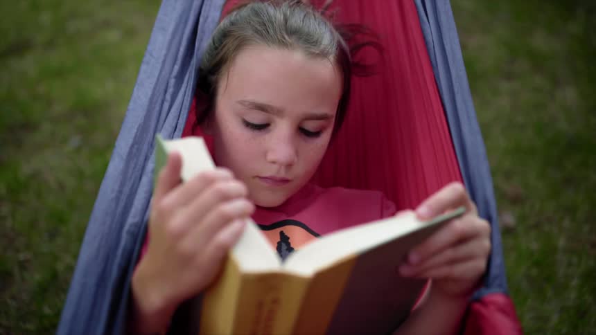 Girl in a hammock reads a book in the backyard