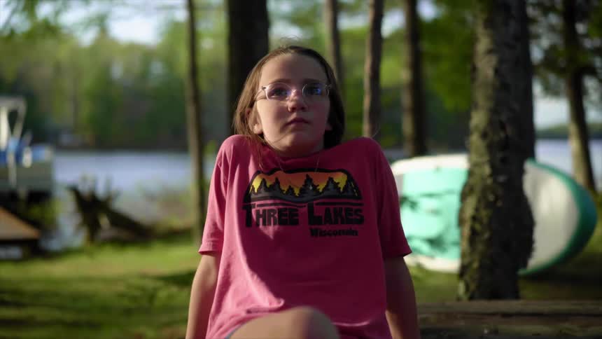 A young girl wearing eyeglasses sits on an old wooden table near a river where a boat is docked