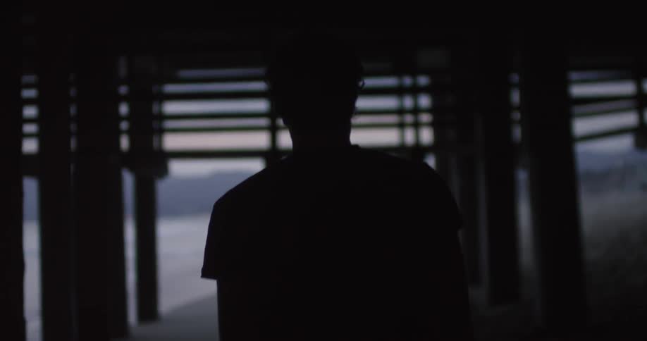 A man walks in the shadows underneath a pier on a beach at dusk