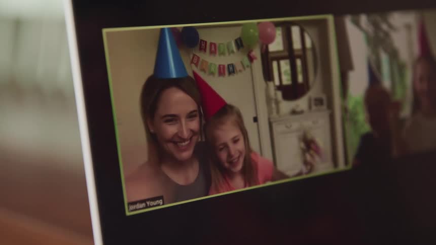 Mother and daughter wearing party hats wave and smile on the screen of a laptop