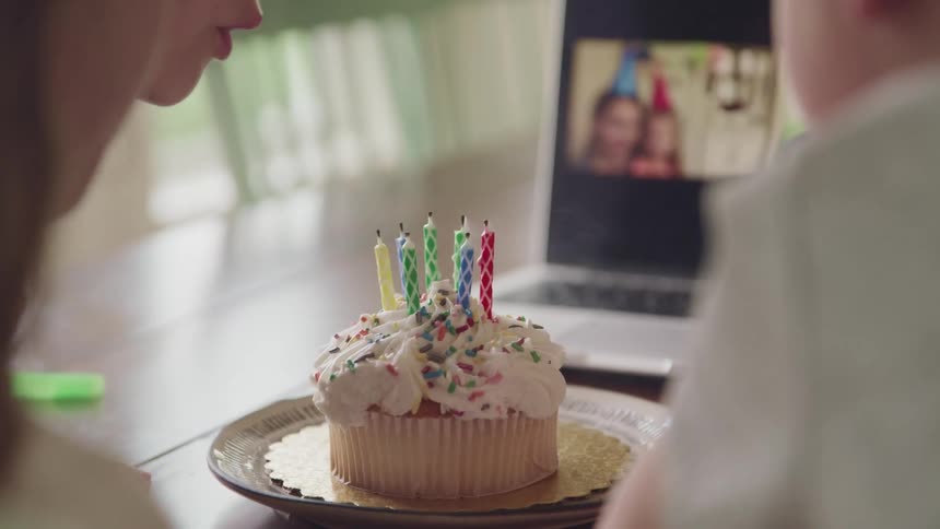 Child blows out the candles on a birthday cake as people watch on a computer