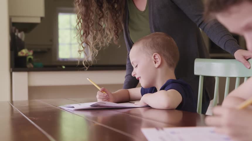 Two boys sit at a table and work as their mother talks with them