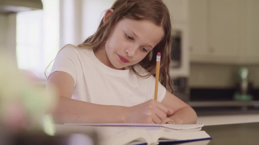 Girl with a white shirt sitting at the table writes in a notebook in the kitchen