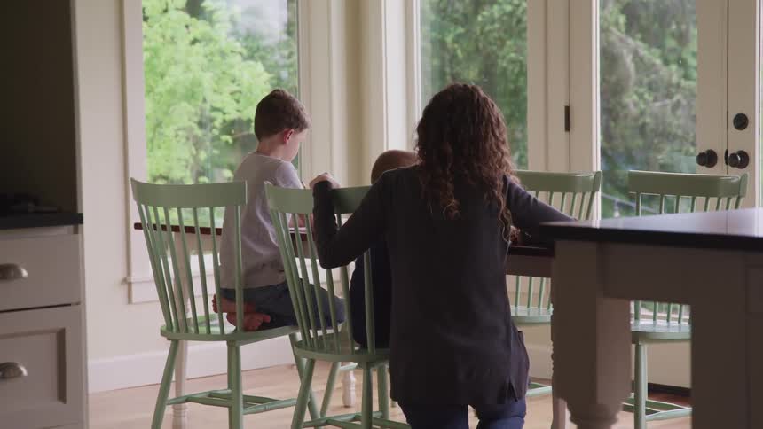 A woman knees next to a table as her sons sit at it