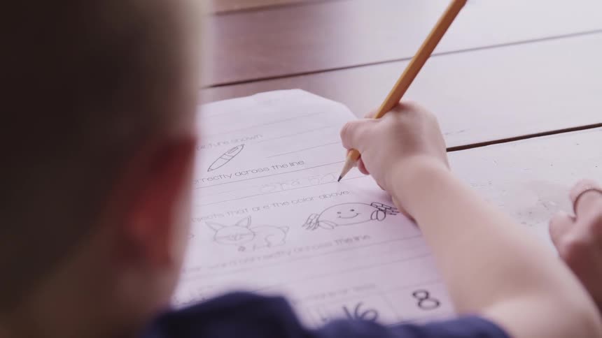 A young boy works on a school assignment while sitting at a table with his mom