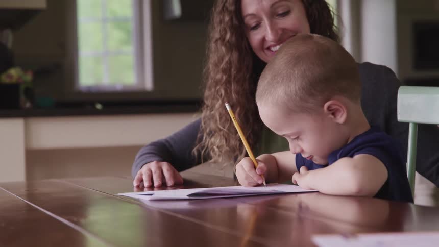 A woman and a boy sitting at a table as the boy writes on a paper with a pencil