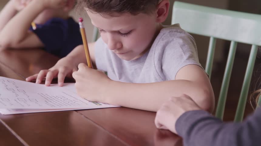 A woman and two boys sitting at a table as a boy writes on the paper with a pencil