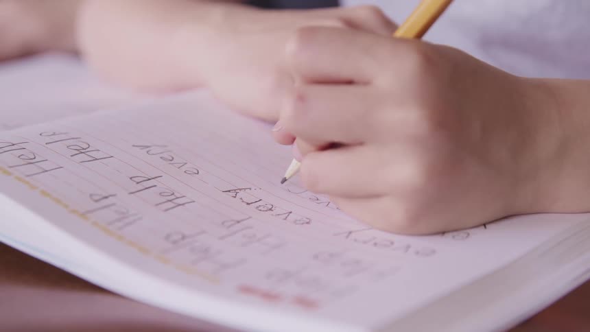 Child with a white shirt sitting at a desk writes with a pencil in a notebook