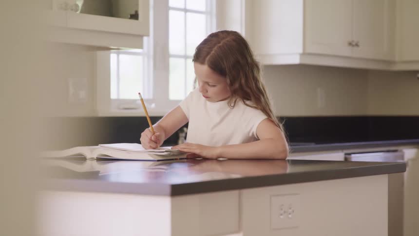 Young girl writes with a pencil in a book while standing in the kitchen