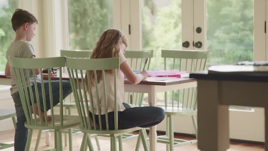 A girl and a boy sit at t a table in their home and draw and color