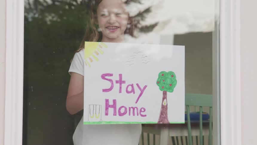 A girl holds a drawing which reads stay home behind the window in her house
