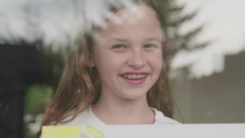 A girl with braces holds a drawing while smiling at the camera through a window