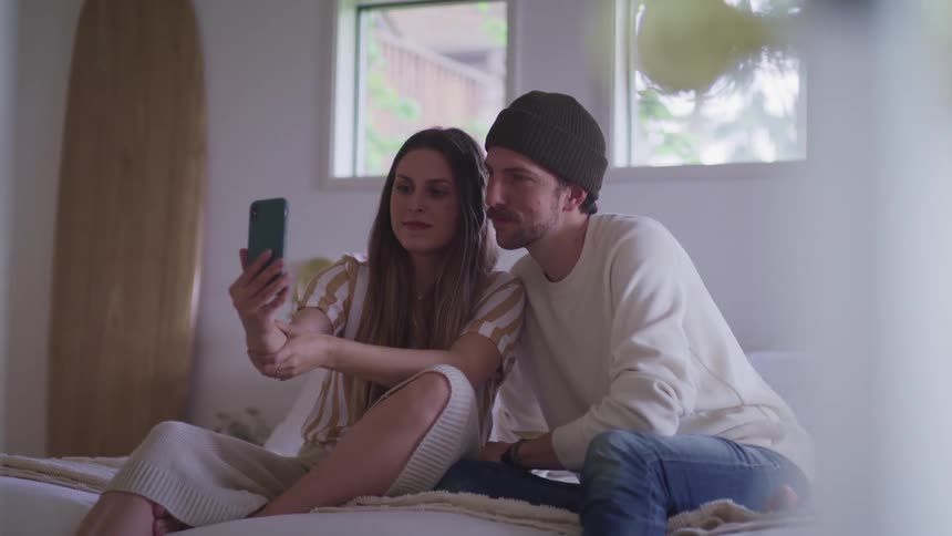 Woman with a striped shirt and man wearing a beanie white sweater and jeans sit on a bed and take a selfie with a smart phone in the bedroom