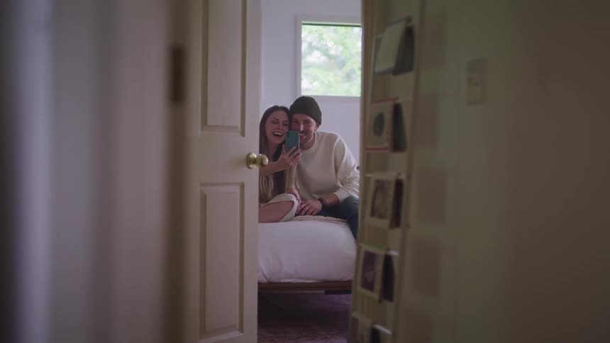 Couple sit on bed laughing and video calling via mobile phone