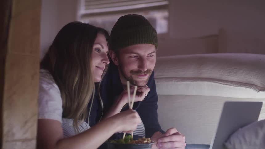 A man and woman lay in bed eating food while watching something on a laptop
