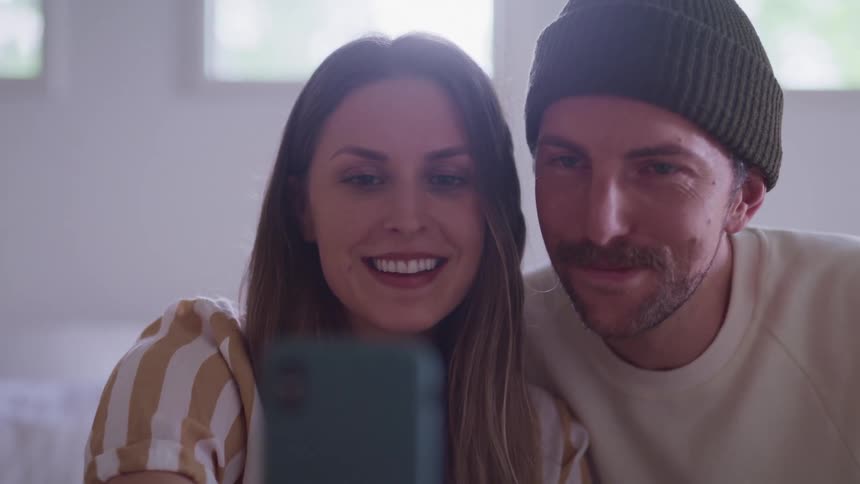 Woman with a striped shirt and brown hair and man wearing a dark green beanie and a white sweater sitting on the bed make a video call on a smart phone in the bedroom during daylight