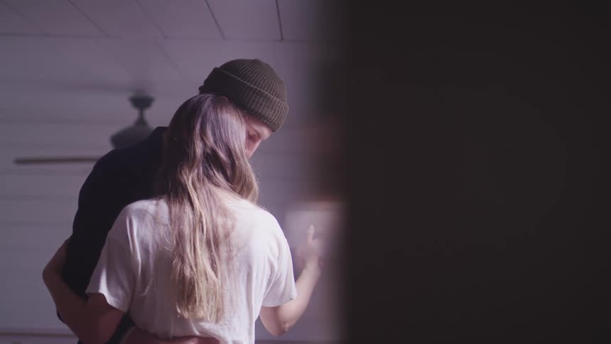 Man with a dark blue shirt and a brown beanie and woman with long brown hair and a white shirt dance together in a room